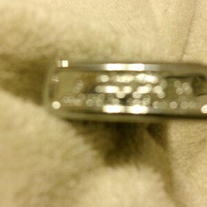 May include: A silver-colored ring with blurred text. The ring is rectangular and appears to be made of metal. The ring is resting on a soft, off-white surface. The text on the ring is not clear.