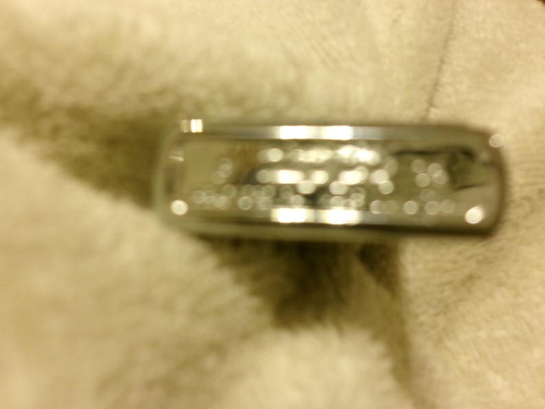 May include: A silver-colored ring with blurred text. The ring is rectangular and appears to be made of metal. The ring is resting on a soft, off-white surface. The text on the ring is not clear.