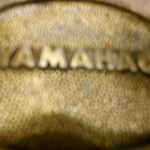 May include: Close-up of a textured, golden-colored surface with the embossed word "YAMAHA" in silver. The surface has a dimpled pattern, and the letters are slightly blurred.