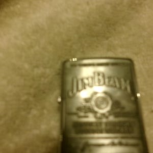 May include: A silver-colored lighter with the "Jim Beam" logo prominently displayed. The lighter has a rectangular shape and appears to be made of metal. The logo is in a stylized font, and the overall design suggests a classic, vintage style.