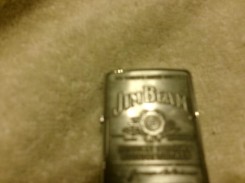 May include: A silver-colored lighter with the "Jim Beam" logo prominently displayed. The lighter has a rectangular shape and appears to be made of metal. The logo is in a stylized font, and the overall design suggests a classic, vintage style.