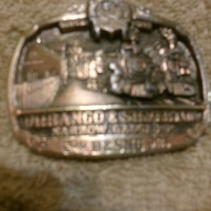 Puede incluir: Una hebilla de cinturón de plata con una imagen grabada de un tren y el texto "Durango & Silverton Narrow Gauge RR" y "Silverton, CO".