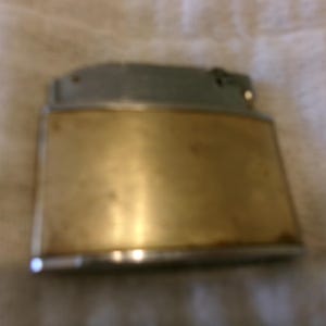 Puede incluir: Un encendedor vintage con cuerpo rectangular de lat&oacute;n y parte superior plateada. El encendedor muestra signos de desgaste, lo que sugiere que es una antig&uuml;edad o un art&iacute;culo muy usado. El lat&oacute;n tiene un tono dorado c&aacute;lido.
