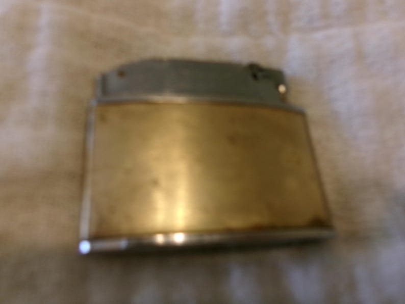 Puede incluir: Un encendedor vintage con cuerpo rectangular de lat&oacute;n y parte superior plateada. El encendedor muestra signos de desgaste, lo que sugiere que es una antig&uuml;edad o un art&iacute;culo muy usado. El lat&oacute;n tiene un tono dorado c&aacute;lido.