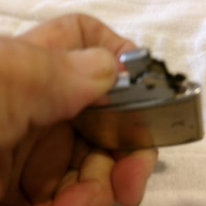 Puede incluir: Un encendedor met&aacute;lico vintage con forma rectangular redondeada. El encendedor tiene una parte superior plateada y un cuerpo dorado. El mecanismo es visible y el encendedor parece estar usado. El encendedor se sostiene en una mano.