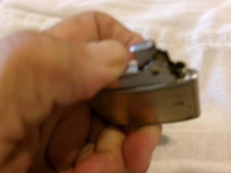 Puede incluir: Un encendedor met&aacute;lico vintage con forma rectangular redondeada. El encendedor tiene una parte superior plateada y un cuerpo dorado. El mecanismo es visible y el encendedor parece estar usado. El encendedor se sostiene en una mano.