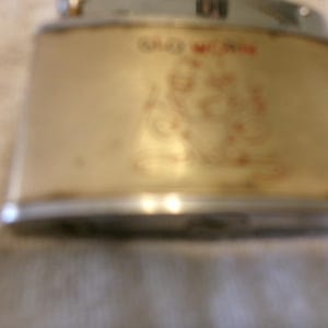 Puede incluir: Encendedor dorado vintage con un dise&ntilde;o rojo y dorado. El encendedor tiene forma rectangular con bordes redondeados y una parte superior plateada. El texto "OLD MONK" es visible en rojo.