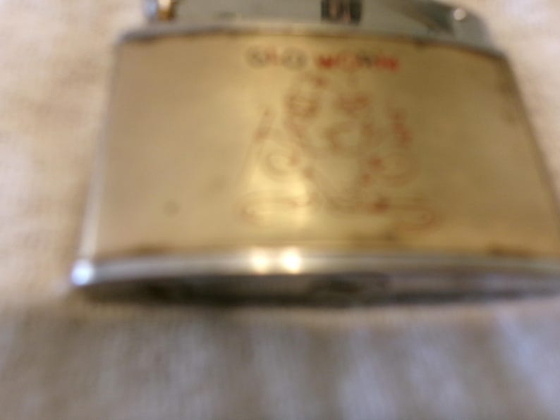 Puede incluir: Encendedor dorado vintage con un dise&ntilde;o rojo y dorado. El encendedor tiene forma rectangular con bordes redondeados y una parte superior plateada. El texto "OLD MONK" es visible en rojo.