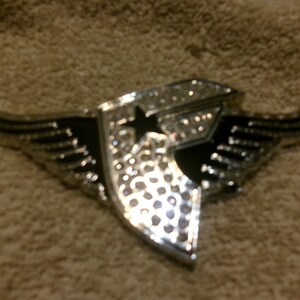 May include: Silver emblem with a star and wing design. The emblem has a central star and is encrusted with small, sparkling embellishments. The wings are detailed with lines, and the emblem has a black background.