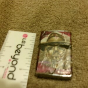 May include: A rectangular lighter with a design featuring a person wearing a hat. The lighter has a silver metal frame with a colorful design. A white ruler with the text "LG beyond" is next to the lighter.