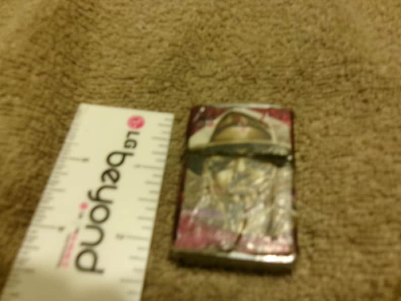 May include: A rectangular lighter with a design featuring a person wearing a hat. The lighter has a silver metal frame with a colorful design. A white ruler with the text "LG beyond" is next to the lighter.