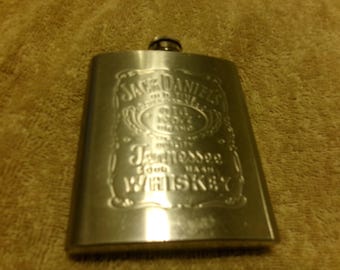 May include: A stainless steel flask with the embossed Jack Daniel's Tennessee Whiskey logo. The rectangular flask has a rounded top and a small, circular cap. The background is a textured, tan surface.