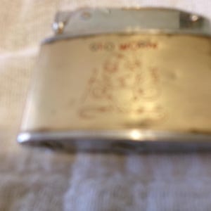 Puede incluir: Encendedor vintage con cuerpo color crema y detalles plateados. El encendedor presenta texto rojo descolorido y un dise&ntilde;o decorativo. El encendedor tiene forma trapezoidal.