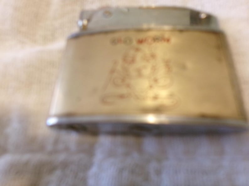 Puede incluir: Encendedor vintage con cuerpo color crema y detalles plateados. El encendedor presenta texto rojo descolorido y un dise&ntilde;o decorativo. El encendedor tiene forma trapezoidal.