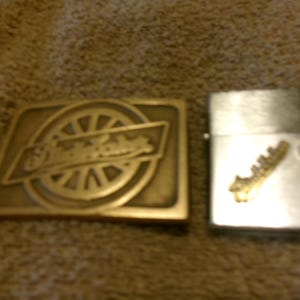 May include: A rectangular brass belt buckle with the text "Olde Tyme" and a wheel design. Next to it is a silver lighter with a gold-colored emblem.
