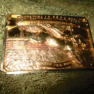 Op de afbeelding: Een gouden metalen gesp met een afbeelding van een paard dat over een hek springt. De gesp heeft de tekst "MONTEZUMA J.C. P.R.C.A. RODEO" en "AUGUST 23, 1969" erop.