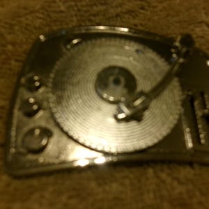 May include: A silver-colored, vintage-style turntable. The design includes a circular record, a tonearm, and control knobs. The overall shape is rectangular with rounded corners, suggesting a retro aesthetic.