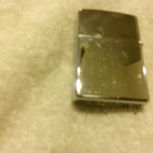 May include: A shiny, silver metal lighter with a rectangular shape. The lighter has a hinged lid and a smooth, reflective surface. The lighter is slightly scratched and shows signs of use.
