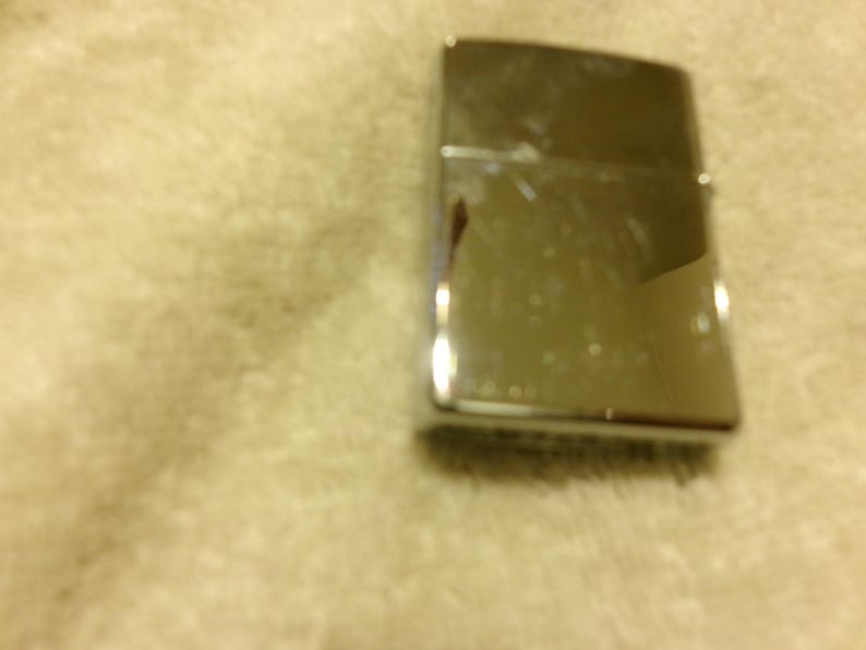May include: A shiny, silver metal lighter with a rectangular shape. The lighter has a hinged lid and a smooth, reflective surface. The lighter is slightly scratched and shows signs of use.