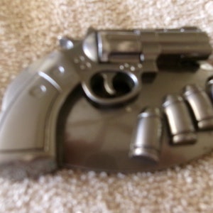 Könnte beinhalten: Eine silberne Gürtelschnalle in Form eines Revolvers mit Lauf und Griff. Die Schnalle hat fünf kugelförmige Akzente an der Seite.