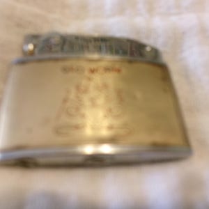 Puede incluir: Un encendedor vintage con forma rectangular y parte superior plateada. El encendedor tiene un cuerpo color crema con texto y elementos de dise&ntilde;o rojos. El texto "OLD W. WIN" es visible en la parte delantera.