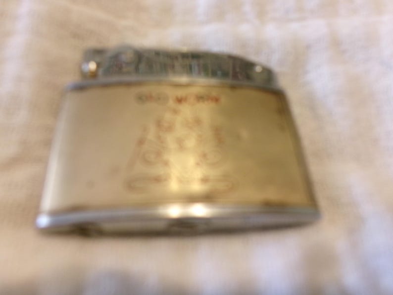 Puede incluir: Un encendedor vintage con forma rectangular y parte superior plateada. El encendedor tiene un cuerpo color crema con texto y elementos de dise&ntilde;o rojos. El texto "OLD W. WIN" es visible en la parte delantera.