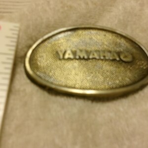 May include: Oval-shaped, brass-colored belt buckle with the word "YAMAHA" embossed on the front. The buckle has a textured surface and a silver-colored border. A ruler is visible on the left side.