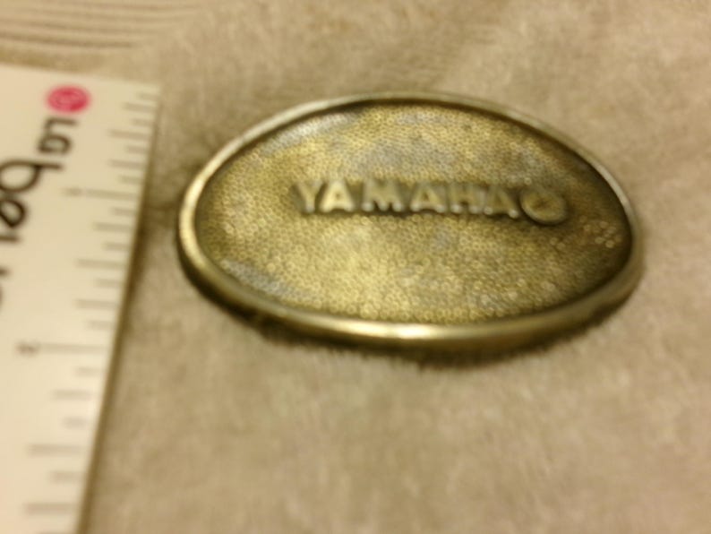 May include: Oval-shaped, brass-colored belt buckle with the word "YAMAHA" embossed on the front. The buckle has a textured surface and a silver-colored border. A ruler is visible on the left side.