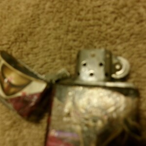 May include: A silver metal lighter with an open lid, revealing a patterned interior. The lighter's body has an embossed design. The background is a blurred, textured brown surface.