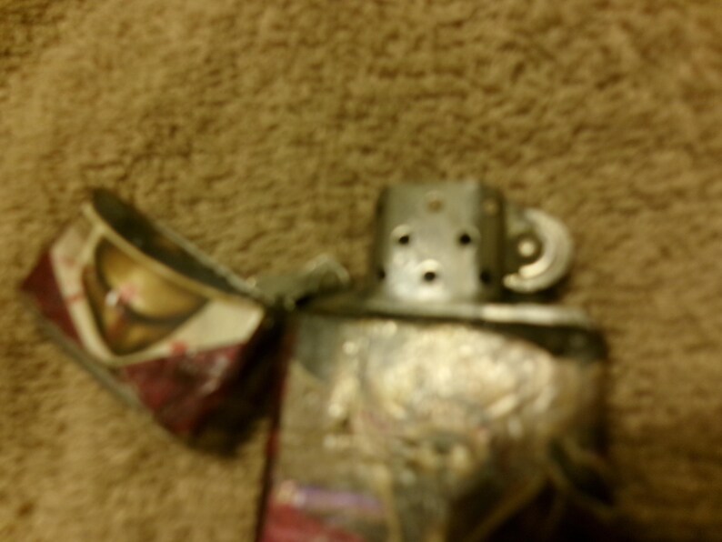 May include: A silver metal lighter with an open lid, revealing a patterned interior. The lighter's body has an embossed design. The background is a blurred, textured brown surface.