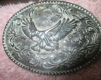 May include: Silver-toned oval belt buckle with an embossed design. The buckle features a central eagle with outstretched wings, surrounded by floral and scrollwork patterns. The edge has a beaded border.