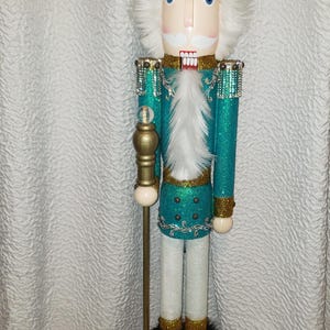 May include: A teal and gold glitter nutcracker with a white beard and a crown. The nutcracker is holding a gold staff with a clear crystal ball on top. The nutcracker is standing on a red base with gold trim.