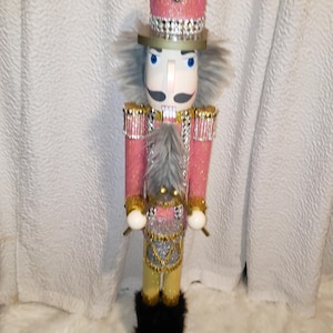 May include: A pink and gold glitter nutcracker with a black fur trim and rhinestones. The nutcracker is holding a gold drum and has a white face with blue eyes.
