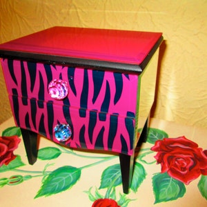 May include: A small pink and black zebra print wooden jewelry box with two drawers and clear crystal knobs. The box is sitting on a table with a painted floral design.