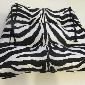 May include: A square seat cushion featuring a black and white zebra stripe pattern. The cushion is tufted with three distinct horizontal sections and includes black fabric ties at the top corners for securing the item to a chair or furniture frame.