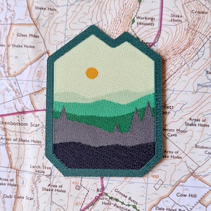May include: A green, yellow, and gray embroidered patch with a mountain range and a sun in the sky. The patch is shaped like a house and has a dark green border.