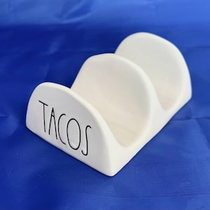 May include: A white ceramic taco holder with three slots. The word "TACOS" is written in black script on the front. The holder is designed to keep tacos upright and is perfect for serving.