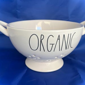 May include: A white ceramic bowl with two handles and the word "ORGANIC" in black lettering. The bowl has small holes around the base and sits on a small pedestal. The background is a solid blue.