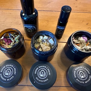 May include: An assortment of apothecary items, including black glass bottles and jars with lids. The jars contain dried herbs and flowers. The lids are labeled with text that reads "Hedge Hearth & Heart" and product names like "Goddess Blend".