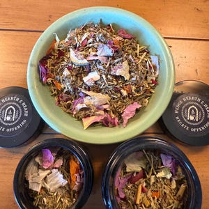 Puede incluir: Un cuenco verde claro y dos pequeños frascos negros llenos de hierbas secas y pétalos de flores. El cuenco está lleno de una mezcla de hierbas secas y pétalos de flores rosas y blancos. Dos pequeños frascos negros están llenos de hierbas y pétalos similares.