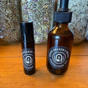 May include: Two brown glass bottles with black caps and labels that read "Hedge Hearth & Heart Protection Oil." One is a roll-on bottle, the other has a dropper. The bottles are on a wooden surface, with jars of herbs in the background.