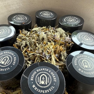 May include: An overhead shot of seven black containers with silver labels, arranged around a pile of dried herbs and flowers. The labels read "Hedge Hearth & Heart Whisperveil" with a graphic of a cauldron. The containers are filled with a product.