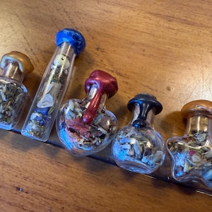Peut inclure: Cinq petits flacons en verre remplis d'herbes et de fleurs séchées. Les flacons ont des formes différentes, dont un carré, un tube et un cœur. Les bouchons sont de couleurs variées, dont l'or, le rouge et le bleu. Les flacons sont disposés sur une surface en bois.