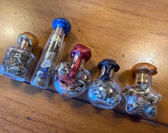 May include: Five small glass bottles filled with dried herbs and flowers. The bottles have different shapes, including a square, a tube, and a heart. The stoppers are various colors, including gold, red, and blue. The bottles are arranged on a wooden surface.