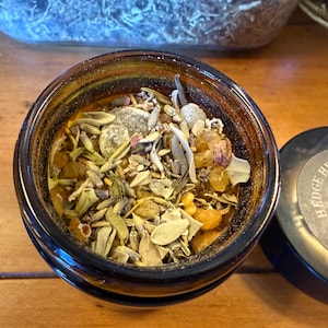 May include: A dark amber glass jar filled with a blend of dried herbs, flowers, and resins. The jar's lid is off to the side, revealing the contents. The lid has the text "Hedge Hearth Angel" in a circular design. The contents include green, yellow, and brown elements.