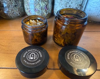 May include: Two small amber glass jars filled with dried herbs and botanicals, with two black lids in the foreground. The lids have a white label with the text "Hedge Hearth & Heart Ancestorcense" around a central illustration.