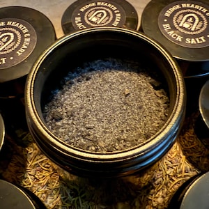 May include: Close-up of a black salt product in a round container. The container is surrounded by other containers with the text "Hedge Hearth & Heart Black Salt". The salt is a dark gray color and appears granular.