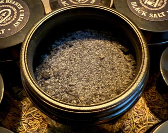 Puede incluir: Primer plano de sal negra en un recipiente redondo. El recipiente está rodeado de otros recipientes con el texto "Hedge Hearth & Heart Black Salt". La sal es de color gris oscuro y parece granulada.