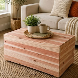 May include: A rectangular wooden coffee table made of light-colored wood with visible grain. The table is placed on a woven rug, with a sofa and decorative items in the background. A small plant, mug, and book rest on a tray atop the table.