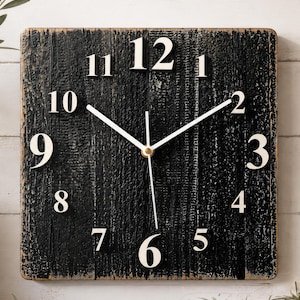 May include: Square black wooden wall clock with white numerals and hands. The clock face has a distressed, textured appearance. The clock hands are white, and the numerals are large and easy to read. The clock is a decorative item.
